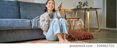Image of young korean woman drinks coffee, holds remote from tv, watching television at home, resting in her house Image of young korean woman drinks coffee, holds remote from tv, watching television at home, resting in her house 109902720
