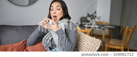 Ill woman using spray from sore throat, treating her cold or flu, staying at home on sick leave, sitting on sofa in living room 109902722