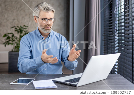 Serious concentrated businessman talking inside office sitting at table with laptop, senior gray haired boss using nuotbook for remote communication online meeting with partners colleagues. 109902737