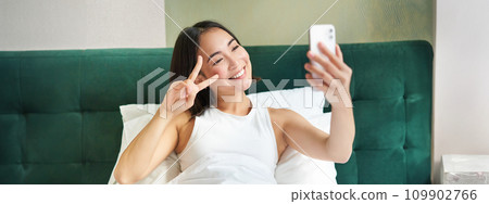 Positive girl blogger takes selfie in her bed, bedroom photo on smartphone, smiling and showing peace v-sign gesture Positive girl blogger takes selfie in her bed, bedroom photo on smartphone, smiling and showing peace v-sign gesture 109902766