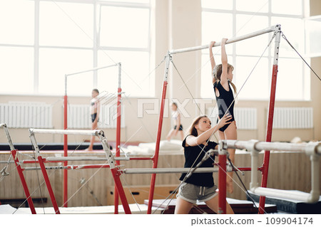 Child gymnastics balance beam. Girl gymnast athlete during an exercise horizontal bar in gymnastics competitions. Coach with child. 109904174