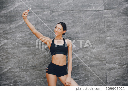 Pretty lady standing in a summer city. Brunette in a black top. Woman near gray wall. Girl with smartphone Pretty lady standing in a summer city. Brunette in a black top. Woman near gray wall. Girl with smartphone 109905021