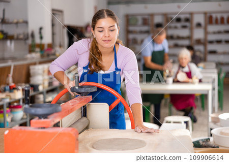 Young woman rolls out clay on craft machine clay press roller in studio 109906614