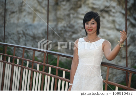 Portrait photography of a mature, beautiful, happy Asian woman. Model in white casual clothes. Standing and smiling at the camera, standing on a bridge with mountains behind. 109910482
