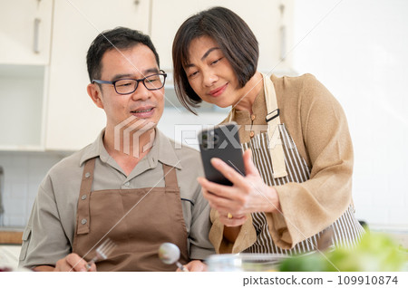 Happy Asian wife is showing something on her phone to her husband while he is eating in the kitchen. 109910874