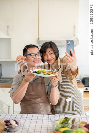 A lovely Asian couple taking pictures or selfies with their food in the kitchen together. A lovely Asian couple taking pictures or selfies with their food in the kitchen together. 109910878