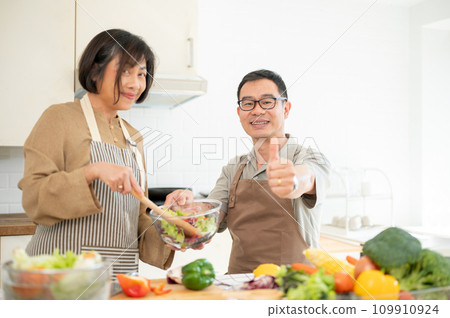 A happy adult Asian husband is showing his thumb up to the camera while enjoying cooking in the kitchen together. 109910924