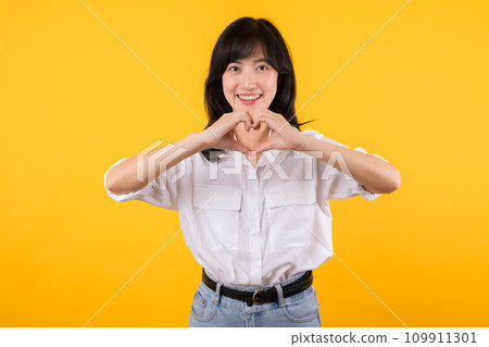 Young female healthcare. Asian woman wearing white shirt feels happy and romantic shapes heart gesture expresses tender feeling poses isolated on yellow background. People affection and care concept. 109911301