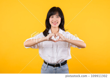 Young female healthcare. Asian woman wearing white shirt feels happy and romantic shapes heart gesture expresses tender feeling poses isolated on yellow background. People affection and care concept. 109911302