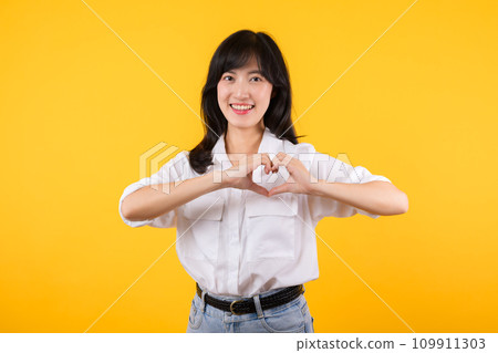 Young female healthcare. Asian woman wearing white shirt feels happy and romantic shapes heart gesture expresses tender feeling poses isolated on yellow background. People affection and care concept. 109911303