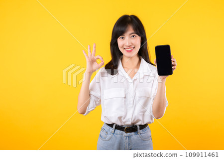 Portrait beautiful young asian woman enterpriser happy smile wearing white shirt and jean plants with okay hand gesture to smartphone isolation on yellow background. new mobile application concept. Portrait beautiful young asian woman enterpriser happy smile wearing white shirt and jean plants with okay hand gesture to smartphone isolation on yellow background. new mobile application concept. 109911461