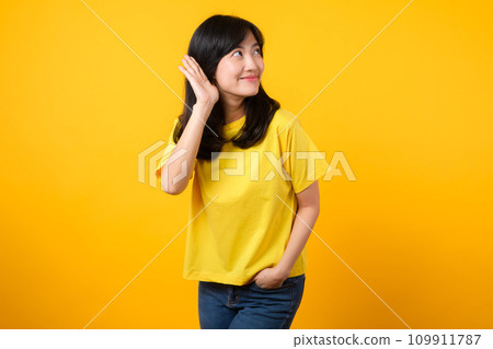 Capture attention portrait of young Asian woman. Wearing a yellow t-shirt and denim jeans, leans in to overhear and listen, evoking curiosity and intrigue. attention-grabbing promotions and discounts. Capture attention portrait of young Asian woman. Wearing a yellow t-shirt and denim jeans, leans in to overhear and listen, evoking curiosity and intrigue. attention-grabbing promotions and discounts. 109911787