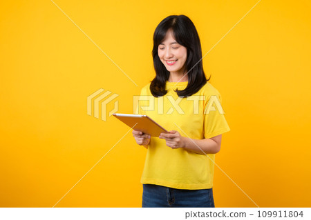 Experience the joy of education technology with portrait. A young Asian woman wearing a yellow t-shirt and denim jeans showcases a happy smile while using digital tablet. education technology concept. Experience the joy of education technology with portrait. A young Asian woman wearing a yellow t-shirt and denim jeans showcases a happy smile while using digital tablet. education technology concept. 109911804