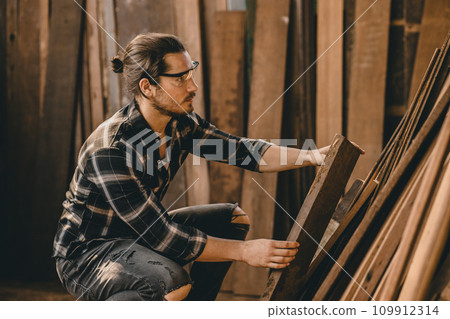 Joiner furniture maker worker choice select wood material from pile stock to find wood piece that suitable for new projects. Joiner furniture maker worker choice select wood material from pile stock to find wood piece that suitable for new projects. 109912314