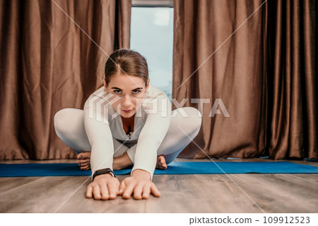 Well looking middle aged woman with long hair, fitness instructor in white sportswear, doing stretching and pilates on yoga mat at home. Healthy lifestyle, harmony, fitness yoga and meditation. 109912523