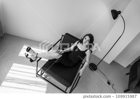 Portrait of a young beautiful girl in a black dress sitting on a chair on a gray background 109915987