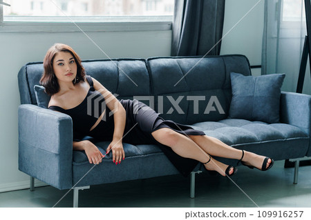 Young beautiful brunette girl in a black dress sitting on the sofa 109916257