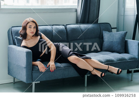 Young beautiful brunette girl in a black dress sitting on the sofa 109916258
