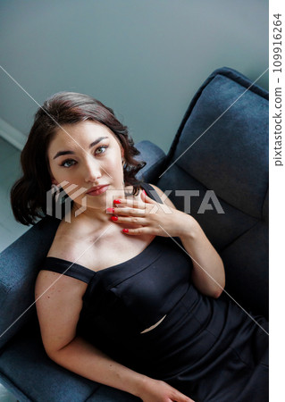 Young beautiful brunette girl in a black dress sitting on the sofa Young beautiful brunette girl in a black dress sitting on the sofa 109916264