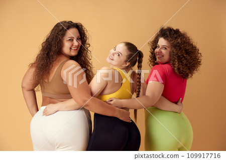 Elegant, positive, smiling young women, friends standing in sportswear over beige studio background. Oversize models 109917716