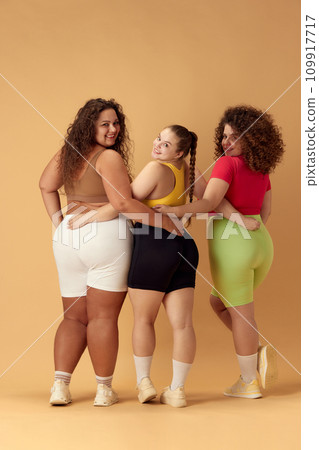 Back view of three smiling young omen, plus size models in comfortable sportswear over beige studio background 109917717