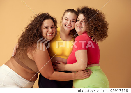 Portrait of three beautiful young women with overweight body standing in sportswear, smiling over beige studio background Portrait of three beautiful young women with overweight body standing in sportswear, smiling over beige studio background 109917718