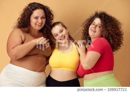 Elegant, positive, smiling young women, friends standing in sportswear over beige studio background. Oversize models Elegant, positive, smiling young women, friends standing in sportswear over beige studio background. Oversize models 109917721