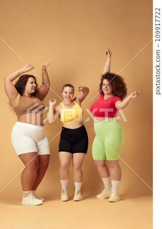 Full-length image of three beautiful women with fat, overweigh bodies in sportswear dancing over beige studio background Full-length image of three beautiful women with fat, overweigh bodies in sportswear dancing over beige studio background 109917722