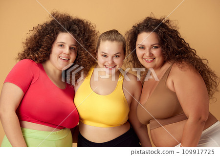 Portrait of three beautiful, smiling, overweigh women standing in sportswear over beige studio background 109917725