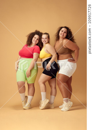 Full-length image of three attractive young women, friends with fat, overweigh bodies standing in sportswear over beige studio background Full-length image of three attractive young women, friends with fat, overweigh bodies standing in sportswear over beige studio background 109917726