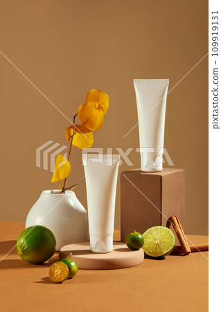 Front view of two cosmetic tubes placed on a wooden platform, fresh lemon, cinnamon and autumn leaves on a brown background. Ideal scene for cosmetics advertising. 109919131