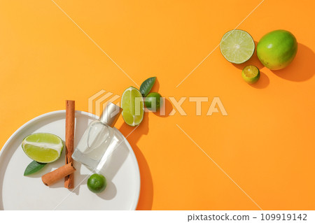 A transparent perfume jar is placed on a white plate with cinnamon, lemon and kumquat on a vibrant background. Mild scent from plants. Copy space for ads. 109919142