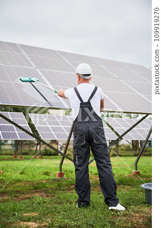 Worker in overalls and helmet cleaning solar battery by mop. Man making sure solar batteries in good condition. Back view of professional cleaning photovoltaic panel. Worker in overalls and helmet cleaning solar battery by mop. Man making sure solar batteries in good condition. Back view of professional cleaning photovoltaic panel. 109919270