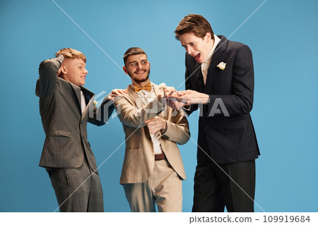 Men in stylish suits emotionally looking on friend showing ring on finger against blue background. Marriage. Shocked faces 109919684