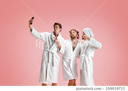 Pretty men in bathrobes taking selfie against pink background. Social media post. Kisses. Cheerful holiday celebration Pretty men in bathrobes taking selfie against pink background. Social media post. Kisses. Cheerful holiday celebration 109919715