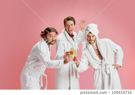 Happy New Year. Positive young men, friends in bathrobes after shower, clinking champagne glasses against pink background Happy New Year. Positive young men, friends in bathrobes after shower, clinking champagne glasses against pink background 109919716