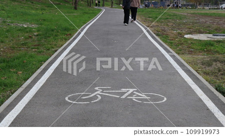 asphalt bicycle path in a park with visible legs of two pedestrians, a bicycle sign painted on the surface of a road in a public urban space, a symbol on the ground asphalt bicycle path in a park with visible legs of two pedestrians, a bicycle sign painted on the surface of a road in a public urban space, a symbol on the ground 109919973