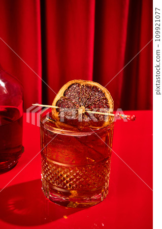 Close-up photo of cocktail, burbon with ice in glass served with dried citrus on toothpick stands on bar counter against red background 109921077