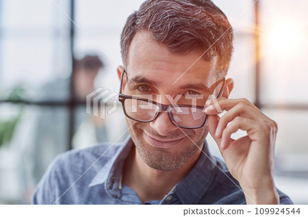 Handsome young guy adjusting his glasses in his office Handsome young guy adjusting his glasses in his office 109924544