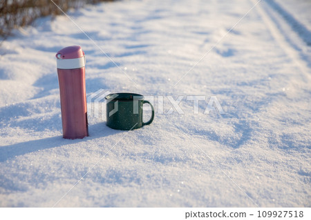 A thermos and a mug standing on a snowy dirt road. 109927518