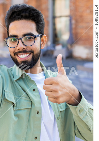 Vertical close-up photo of a smiling young Indian man talking on the phone on a video call, recording a blog, smiling at the camera and showing super. Vertical close-up photo of a smiling young Indian man talking on the phone on a video call, recording a blog, smiling at the camera and showing super. 109928114