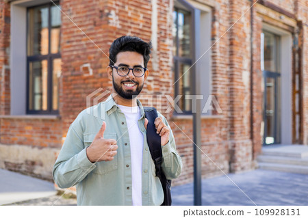 Portrait of a young Indian male student, freelancer standing outside on the street with a backpack, smiling at the camera and showing the super sign with his finger. Portrait of a young Indian male student, freelancer standing outside on the street with a backpack, smiling at the camera and showing the super sign with his finger. 109928131
