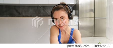 Close up portrait of brunette fitness girl, listens music in wireless headphones, smiles at camera, does workout alone at home, sits on floor and exercises 109928529
