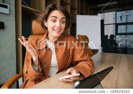 Portrait of woman working in office, looking amazed, makes surprised reaction to big news, holding smartphone, using digital tablet Portrait of woman working in office, looking amazed, makes surprised reaction to big news, holding smartphone, using digital tablet 109928562