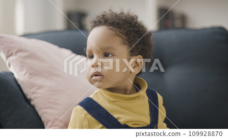 Sweet African American child with natural black hair sitting on sofa and looking around 109928870