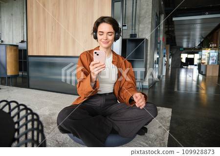 Portrait of stylish young woman employee, digital nomad sitting in office on floor, relaxing in lounge zone, using smartphone and wireless headphones 109928873