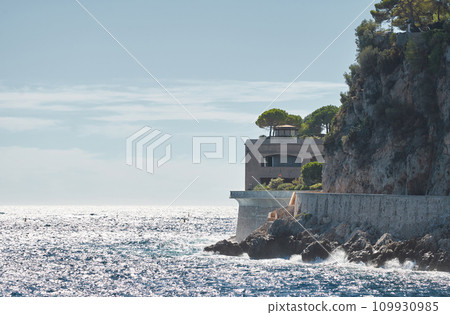 The rocky Promenade of Monaco on a clear sunny day, waves crashing on rocks, green trees on the mountain The rocky Promenade of Monaco on a clear sunny day, waves crashing on rocks, green trees on the mountain 109930985