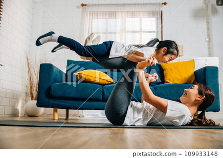 In a cozy living room a mother and her daughter embrace the fun of family yoga pretending to be airplanes in flight. Their bond is strengthened trust is nurtured and happiness is shared. 109933148