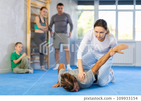 Woman is learning how to grab opponent's hand in prone position during self-defense class, in background girl and boy watch sparring Woman is learning how to grab opponent's hand in prone position during self-defense class, in background girl and boy watch sparring 109933154