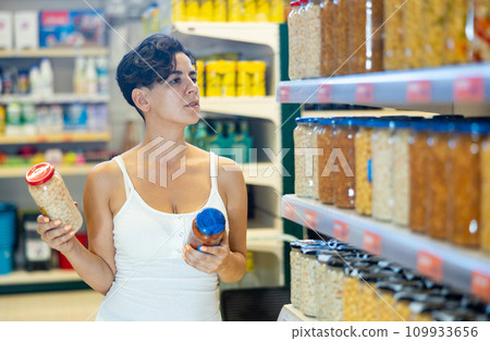 Young Hispanic woman choosing canned beans in glass jars in supermarket Young Hispanic woman choosing canned beans in glass jars in supermarket 109933656